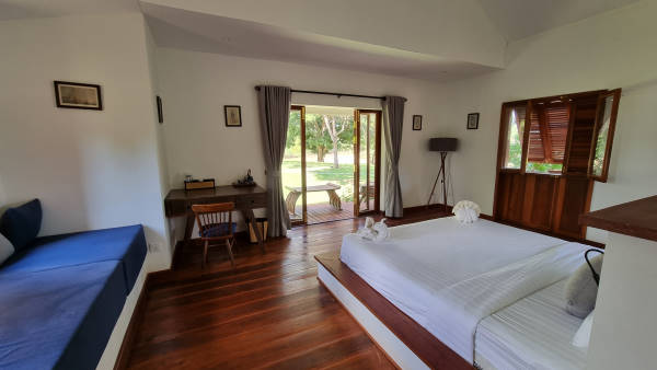 Chambre double avec lit avec des draps blancs face à la baie vitrée donnant sur le jardin et ouverte. A gauche de la fenêtre un bureau en bois