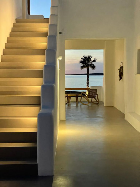 escalier épuré et blanc éclairé par une lumière douce et chaude. Au bout du couloir on voit la mer et un palmier