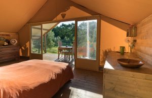Interior of a glamping tent with double bed, wooden furniture and large door opening onto a covered terrace with garden furniture