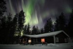 House in the night with the roof covered with snow, the moon lights on and above the fir trees a beautiful aurora borealis