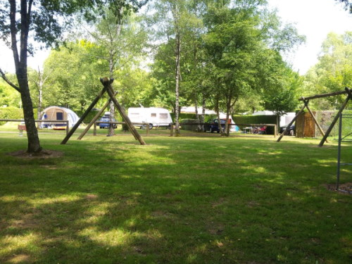 Aire de jeu du camping sous les arbres avec des balançoires en bois. Au fond on voit des caravanes et une tente