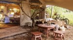 Photo prise sur la terrasse couverte d'une des tentes de glamping. On voit une table basse avec tabouret en bois, un espace pour faire une sieste et au fond la chambre avec la baignoire ancienne au milieu de la pièce.