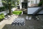 Photo de la cours de l'hotel ensoleillée : murs blancs, petit espace d'herbe, mobilier en fer noir et blanc et grand echiquier au sol avec ses grands pions