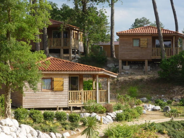 Natureo Seignosse. Photo des 3 chalets en bois construits sur pilotis sur une pente arborée de pins.Ils sont reliés par un chemin de terre bordé de cailloux blanc.