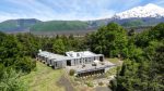 Vue du ciel : les batiments en bois du lodge au milieu d'une clairière entourée d'arbre et au fond les montagnes dont un sommet enneigé