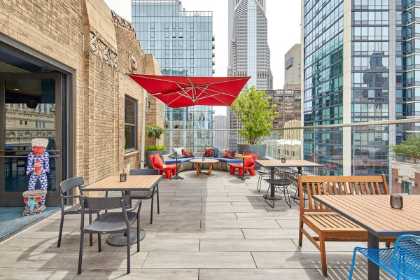 Virgin Hotel Chicago - photo de la terrasse amenagée sur le toit de l'immeubme : un grand canapé gris protégé avec une voile rouge, un table pour 2 et une table plus grande en bois, le tout vue sur les bulding du centre ville