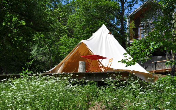 Le bois de Basalte - Tente de glamping posée sur une plateforme au milieu d'une prairie fleurie et entourée d'arbres. derrière on devine un batiment en bois