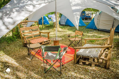 Vegan surf camp - mobilier exterieur (chaise en bois, table bobine, chaise de camping) sur un tapis de sol sous un auvent entouré de tentes