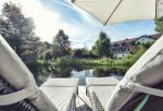 biohotel-schlossgut-oberambach - Photo prise depuis une des rives de l'étang où spnt installés deux transats. De l'autre côté de la rive il y le batiment de l'hotel aux murs blancs et toit en tuiles rouges