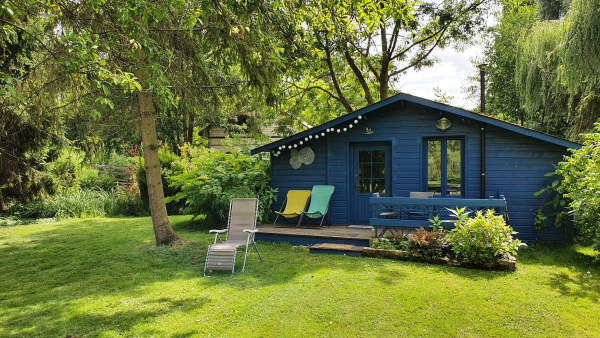 D'une ile a l'autre - Cabane en bois bleu canard avec une girlande luminause sur le toit et un transat jaune et un vert devant sur la terrasse en bois