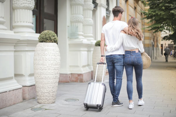 A couple in the street dressed in jeans and a white T-shirt with a small grey wheeled suitcase
