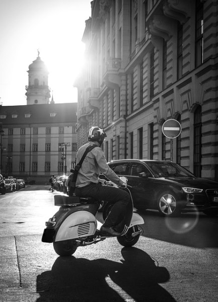 How to get around Italy - Black and white photo of a man on a vintage scooter with a helmet with a star and a shoulder bag in a city center?
