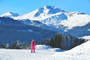 Femme avec une combinaison rose de dos entrain de descendre une piste de ski face à une montagne enneigée. En bas il y a une croix et quelques sapins