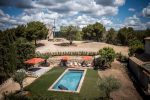 Photo prise avec un frome avec la piscine entourée d'herbe. Autour des transats et des parasols ouverts oranges. Au fond le moulin au milieu des oliviers