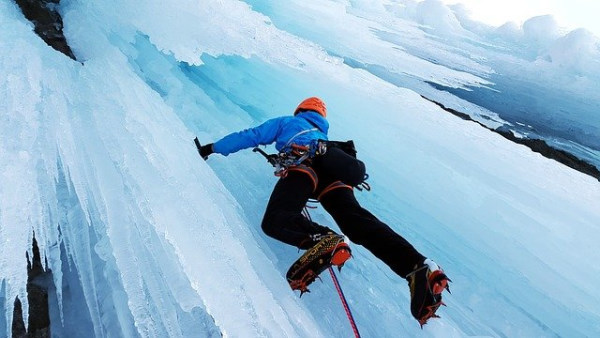 A man wearing a blue coat and an orange helmet is climbing an icefall. He holds an ice axe in his right hand