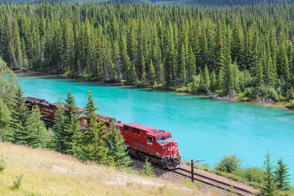 red locomotive coming out of a forest of fir trees. On its right a lake with turquoise waters