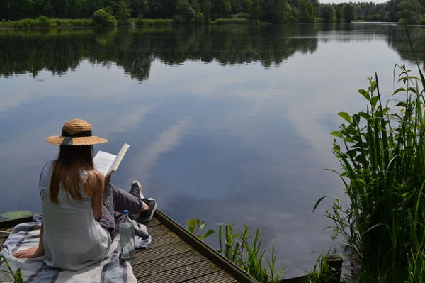 Disconnected vacations : woman with a hat sitting on a pontoon by the water reading a book