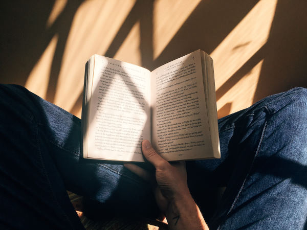 Une personne assis en tailleur sur un sol en parquet couleur claire tient un livre ouvert dans sa main droite