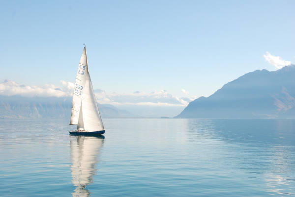 Sailboat with a blue hull and white sails sailing peacefully on a lake surrounded by mountains